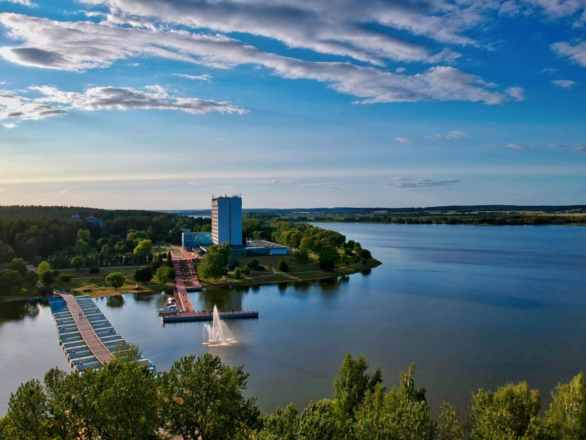 about-img-01 Aerial view of Zaslaŭje, a resort by the lake with a bridge and fountain under a blue sky.
